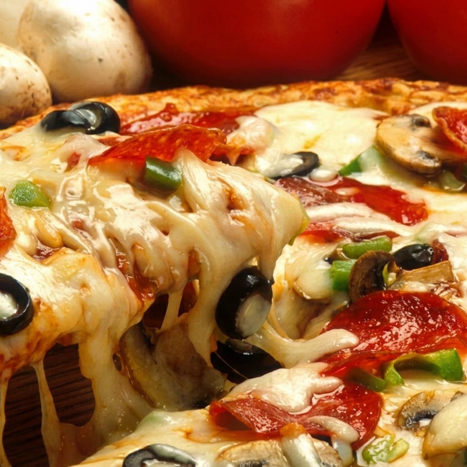 Loaded pepperoni pizza with mushrooms, olives, green peppers, and melted cheese, served with tomatoes and potatoes in the background.